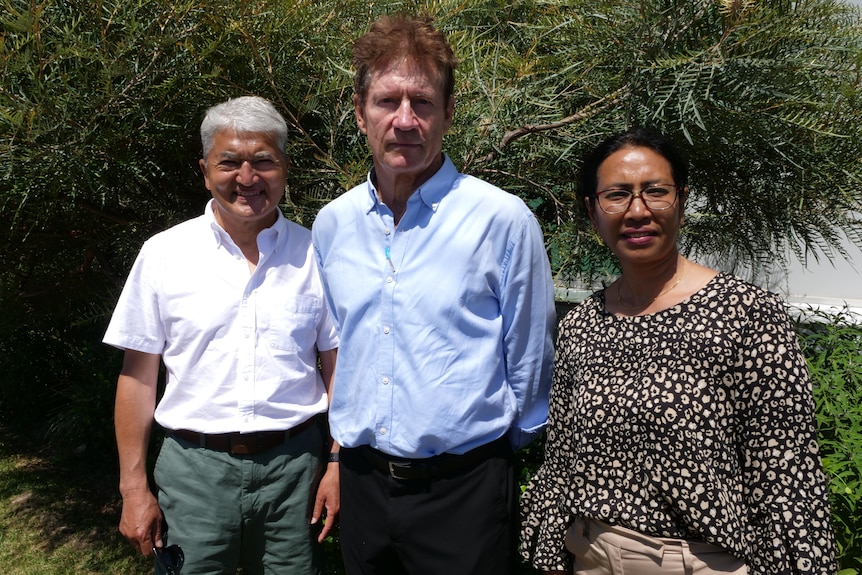 Smiling grey-haired Nepalese man,woman with glasses, either side of  tall Caucasian man, red hair, in front of green bushes.