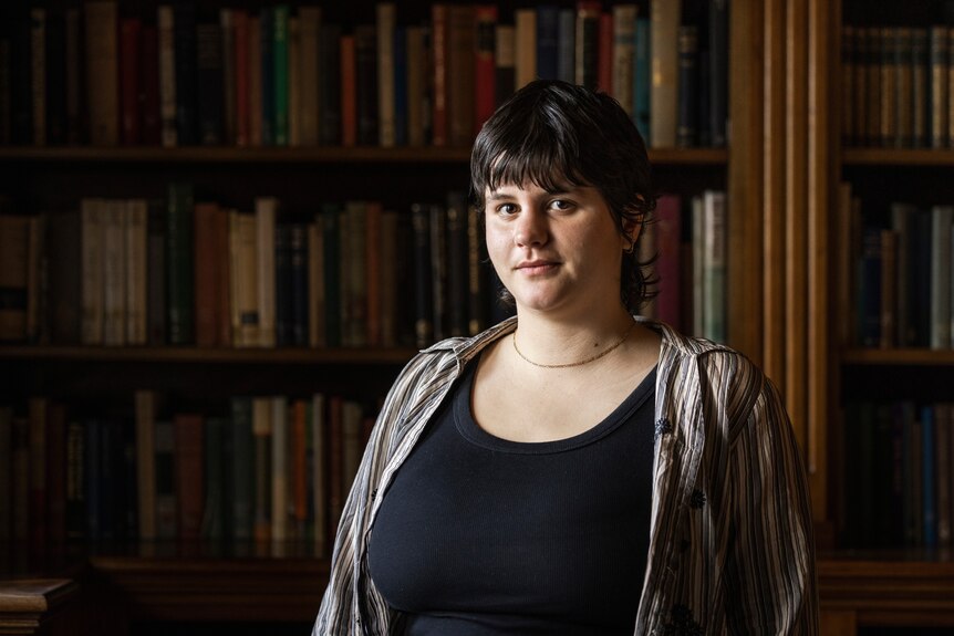A young woman with short dark hair stands in front of library shelves in a dark room, with a gentle smile on her face,