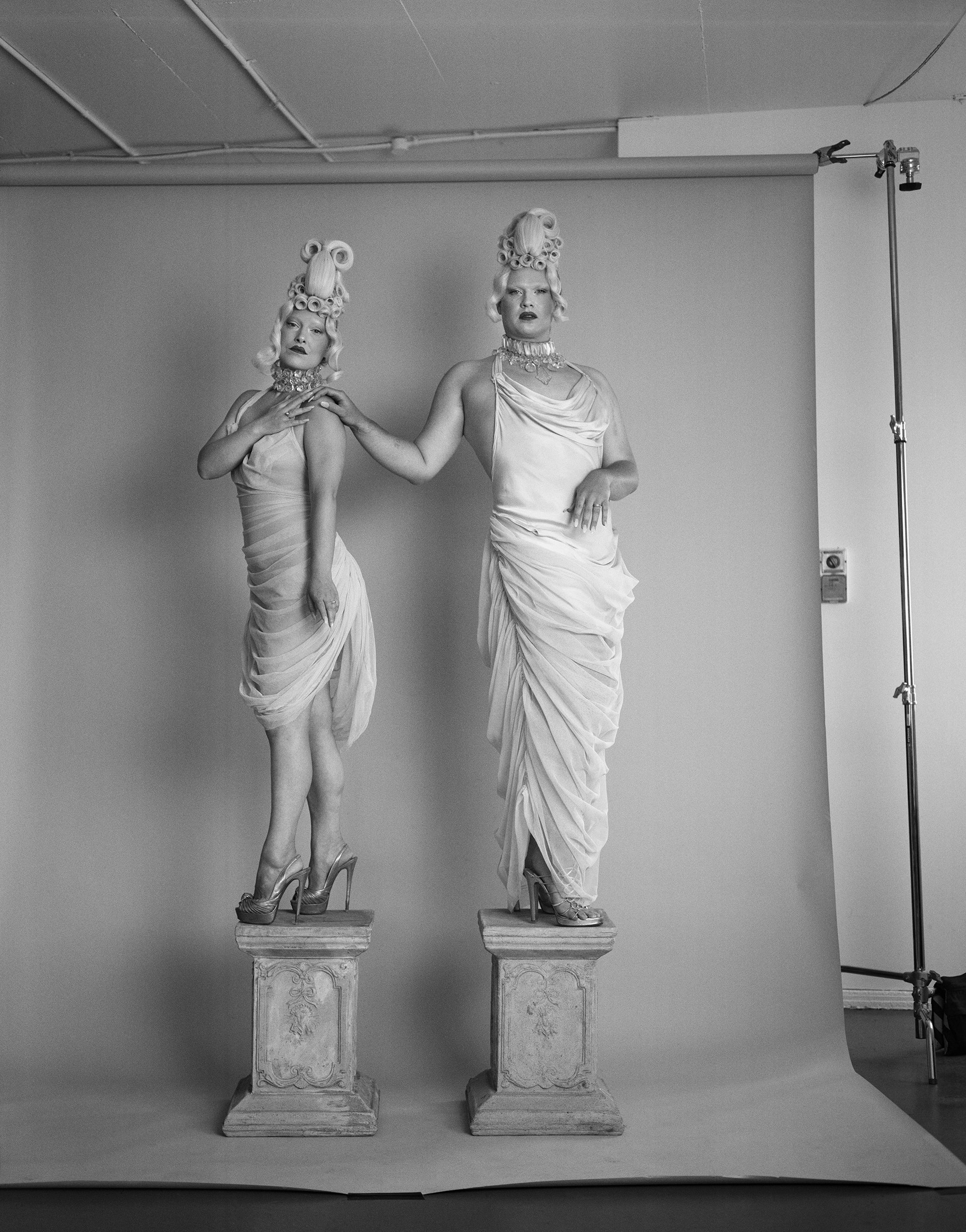 A black and white photo of two women dressed in silk gowns and with elaborate blonde updo hairstyles standing on podiums.