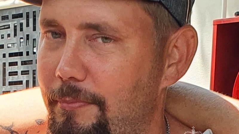 A man with a moustache and goatee and wearing a cap standing in front of a building and smiling. 