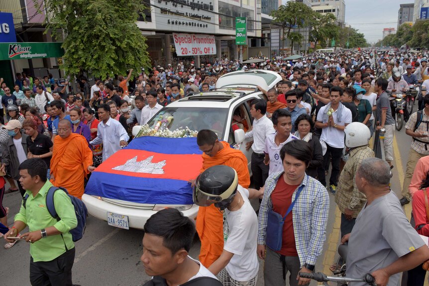 A large crowd fills a street, walking alongside a car covered in flowers and the Cambodian flag.