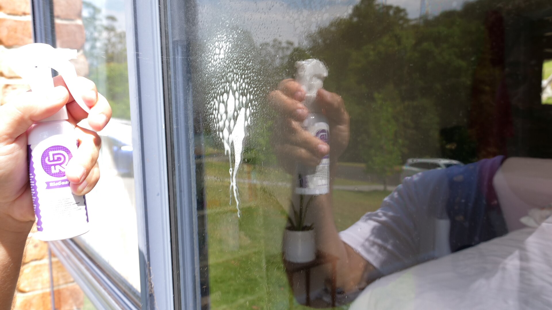 A mão de uma pessoa borrifa um líquido de limpeza na janela de uma casa.