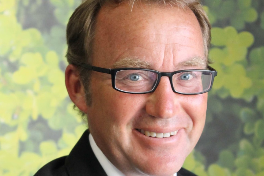 middle aged man with fair hair and black glasses smiling in suit and tie in front of a green floral background