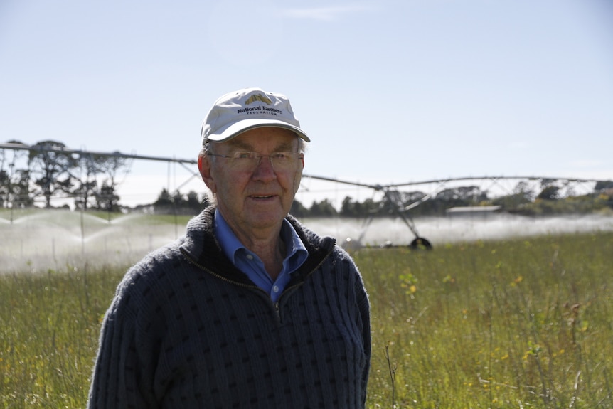 A man in a cap stands in a field with a large pivot irrigator working behind him 
