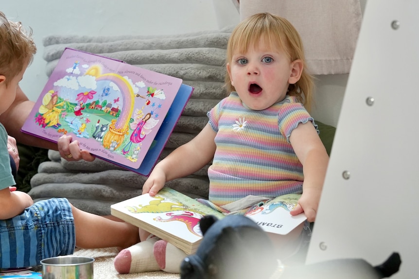 A young white girl with blonde hair yawns while holding a book at daycare in Broken Hill. 