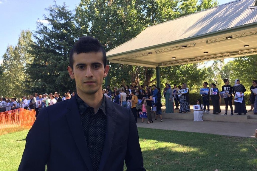 A man with black hair, wearing a dark blazer and collared shirt, stands in front of a group of people holding signs in a park.