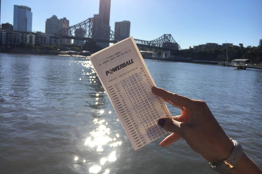 A woman's hand holds up a Powerball ticket in front of the Brisbane River and skyline.