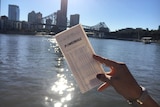 A woman's hand holds up a Powerball ticket in front of the Brisbane River and skyline.