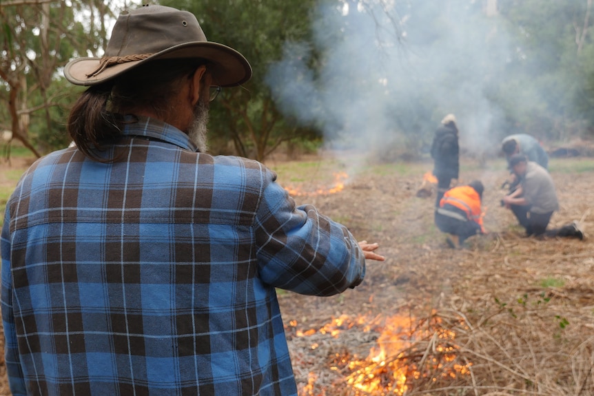 Dave Wandin, in a hat and checked shirt, watches on as students light small "cool" burns.