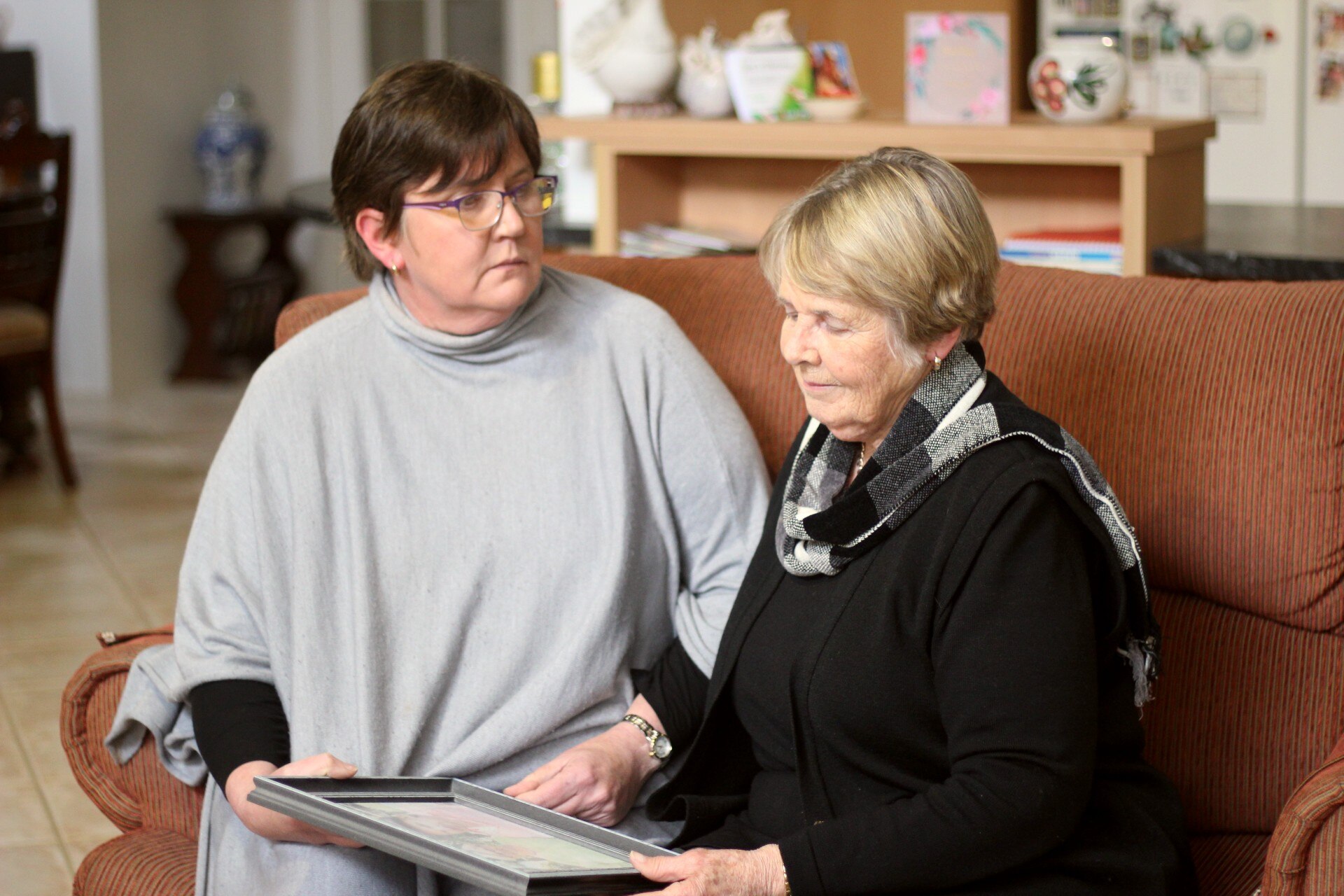 A younger woman in her 50's with a gray poncho and her mother in a gray top, are sitting on a couch looking at a photo