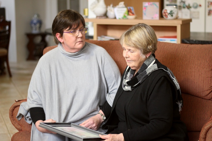 A younger woman in her 50's with a gray poncho and her mother in a gray top, are sitting on a couch looking at a photo