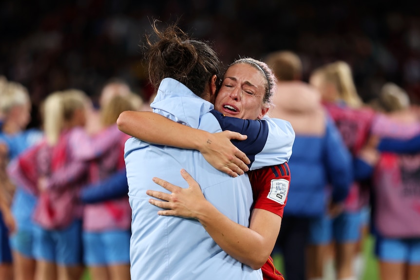 A woman wearing a red shirt cries while hugging someone in a light blue jacket