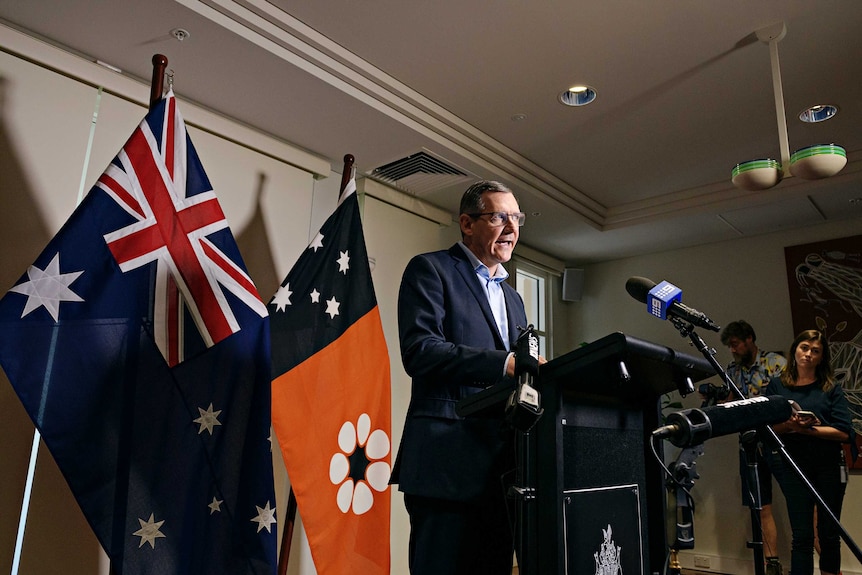 A man giving a press conference at a lecturn with an Australian and NT flag behind him