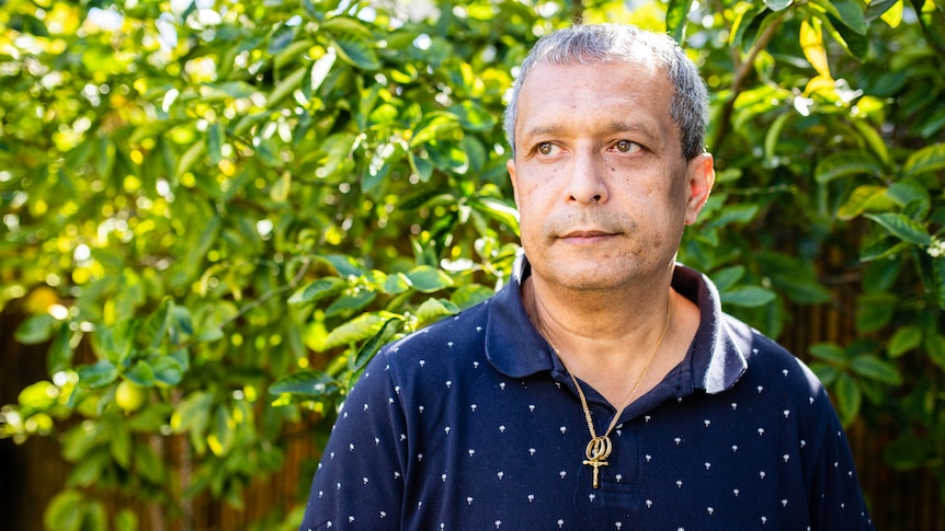 A man in a navy polo shirt looks into the distance, with a tree in the background.