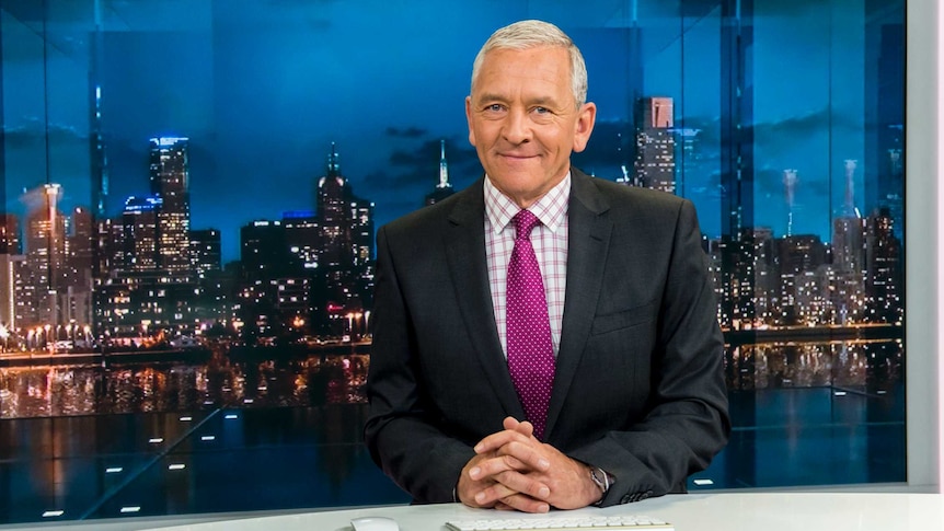 A man in a suit sits on the set of a television news broadcast