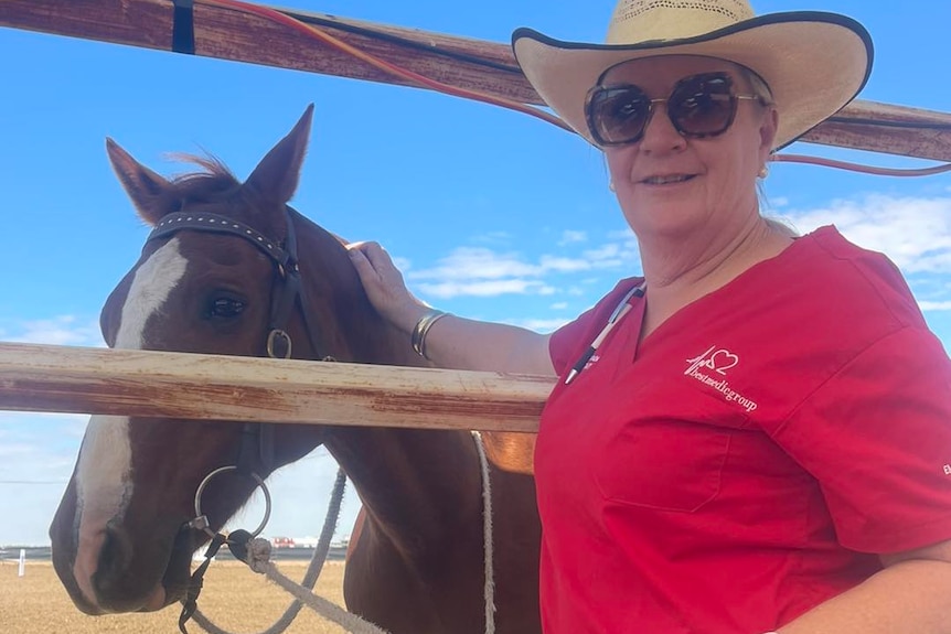 Karen Bath smile at the camera wearing red scrubs, a hat and sunglasses, holding a horse through a rail.