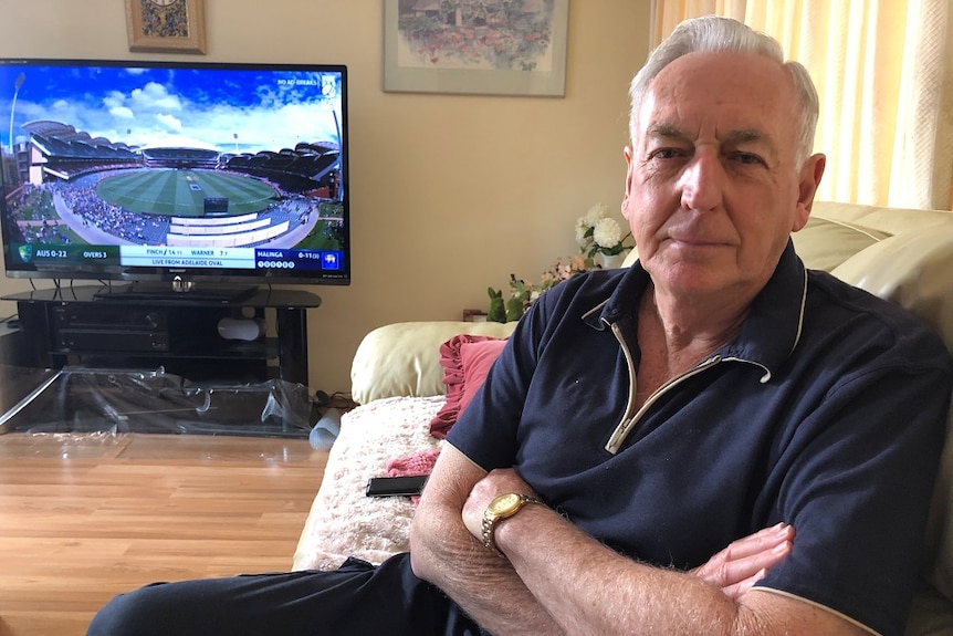 Ron Woodward sits on a couch with his arms crossed looking at the camera next to a television showing Adelaide oval.