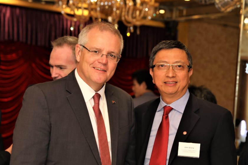 Two men in suits stand in lavish dining room with chandelier and red curtain visible behind.