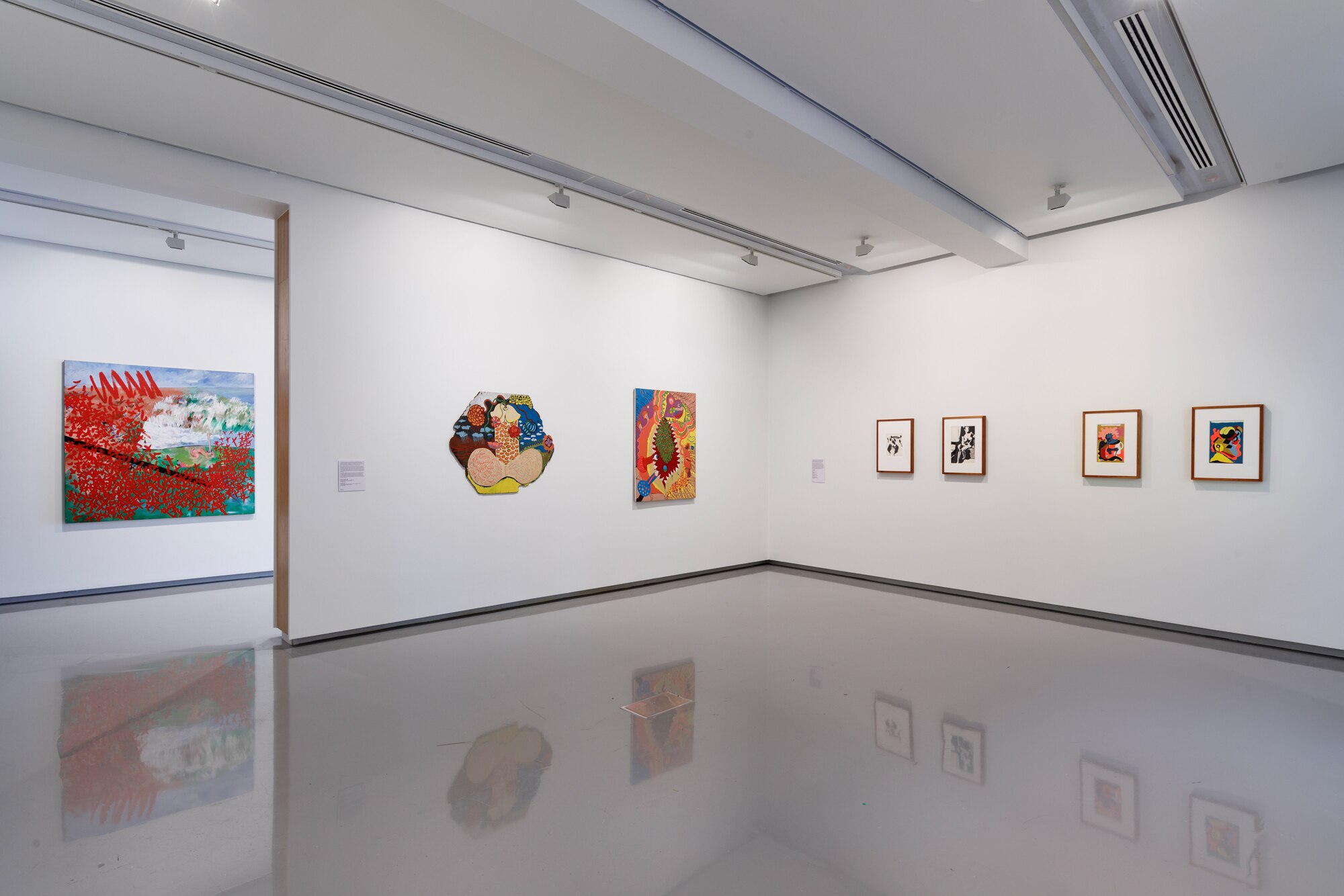 A shot of the interior of a white gallery reveals paintings hung on the wall. The paintings are a riot of colour.