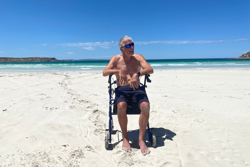 Man with grey hair sitting on a walker at a white sandy beach with clear blue waters in back ground and headland on both sides