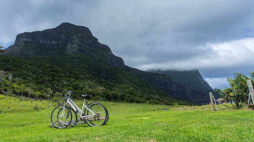 A shadowed Mount Gower rises up into cloud cover, with a green field and a few bicycles in the foreground.