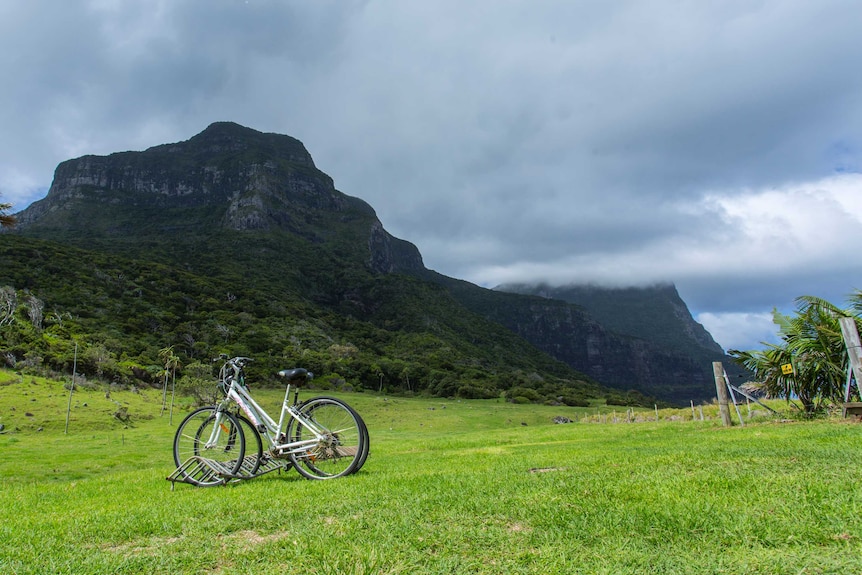A shadowed Mount Gower rises up into cloud cover, with a green field and a few bicycles in the foreground.