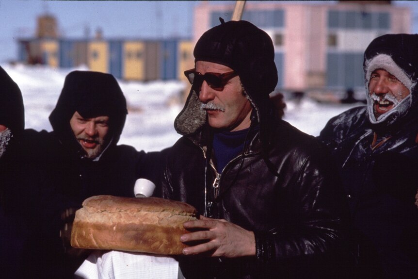 A man holds a loaf of bread as other men stand around in a snowy scene.