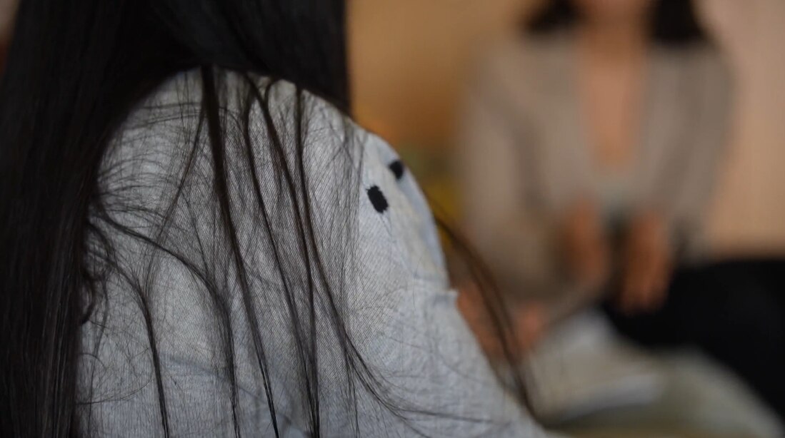 Unidentified woman seated on a couch. She's wearing leggings and a grey cardigan.