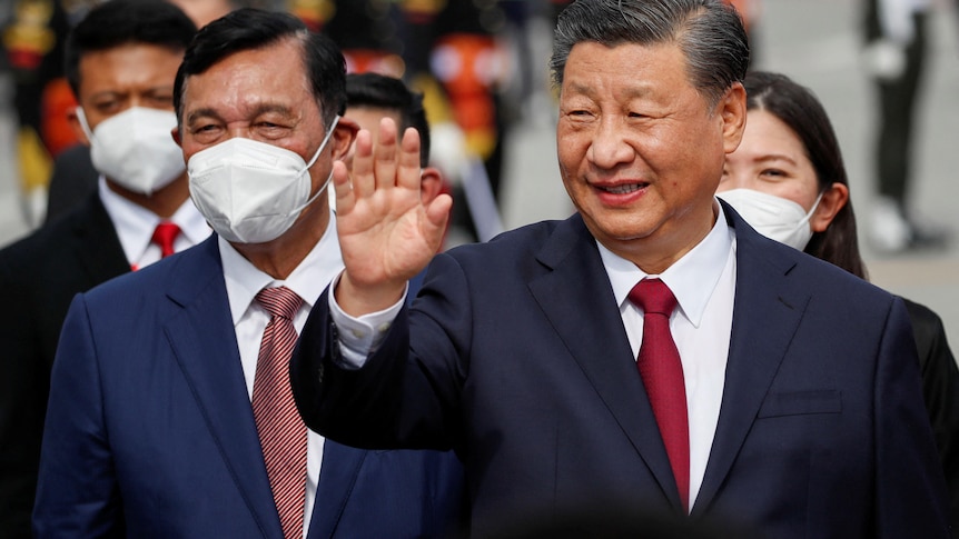 Xi Jinping wearing a suit with red tie waving, with men in masks in the background