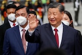 Xi Jinping wearing a suit with red tie waving, with men in masks in the background