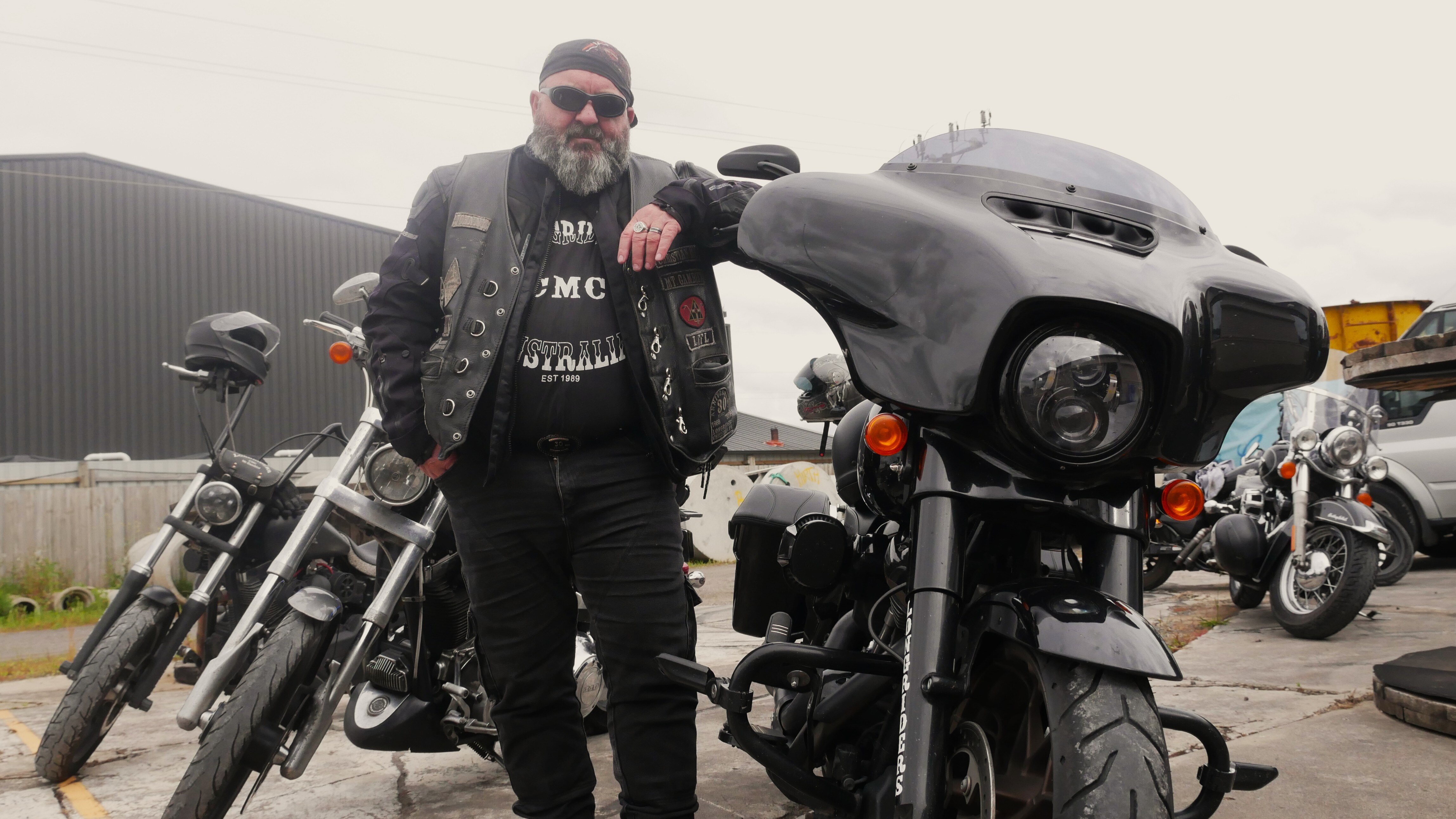 Un hombre vestido con una chaqueta de cuero negra sin mangas, gafas de sol y un sombrero se apoya en el manillar de su motocicleta negra.
