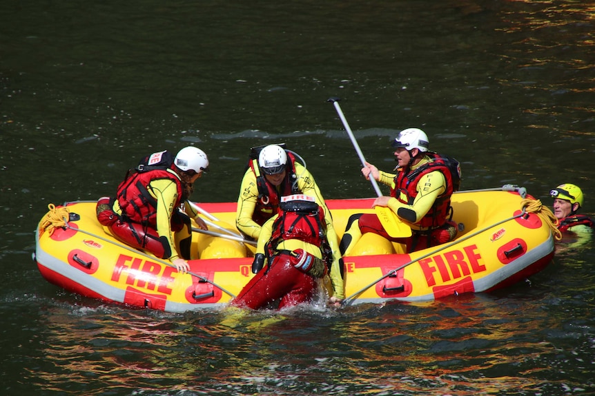 Three people sit in an inflatable boat in diving rescue ppe, one is climbing in from the water