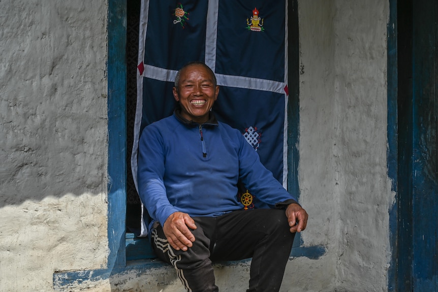 A man in a blue jumper sits on a window ledge in front of an embroidered cloth