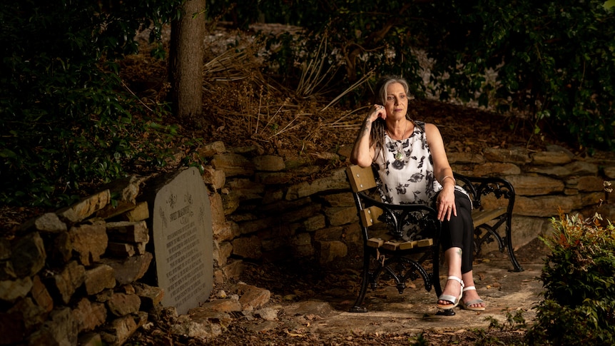 A woman sits on a bench with one hand on her knee and the other resting on her face, next to a memorial plaque