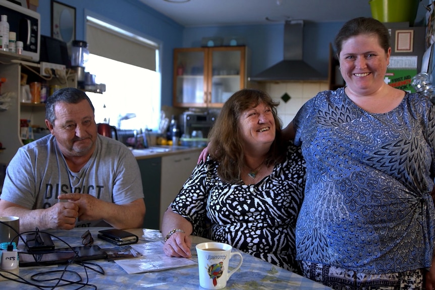 A young woman, smiling at the camera puts her arm around her mum, who is sitting at a table alongside a man.