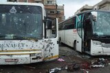 Two buses dotted with bullet holes, blood-soaked streets with shoes lying on the road nearby, buildings in the background