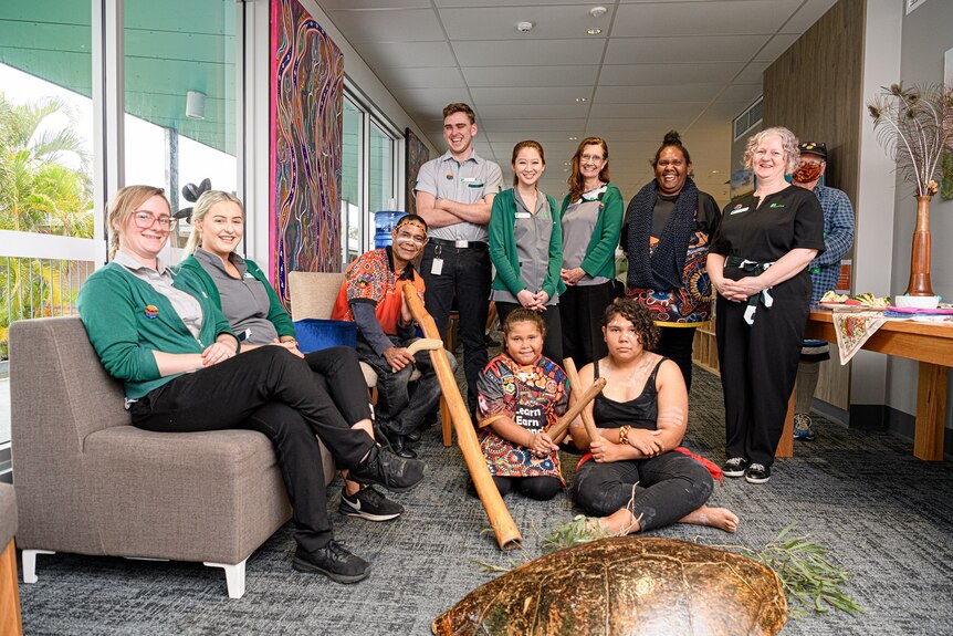 A turtle shell, leaves in the foreground, a team of care staff with local Indigenous artists and dancers in front.