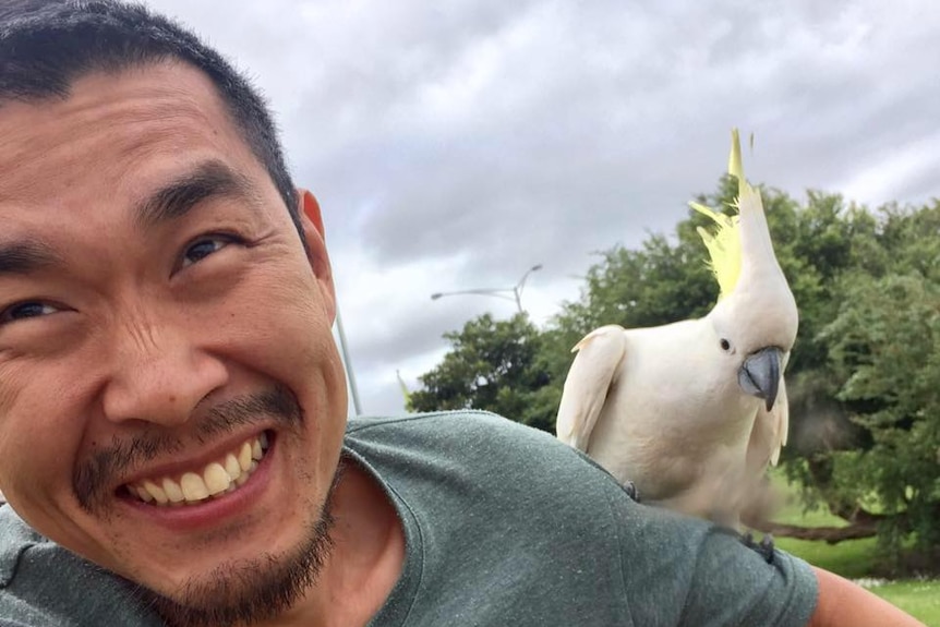 Yun-Seob Shin smiles and sits with a cockatoo on his shoulder.