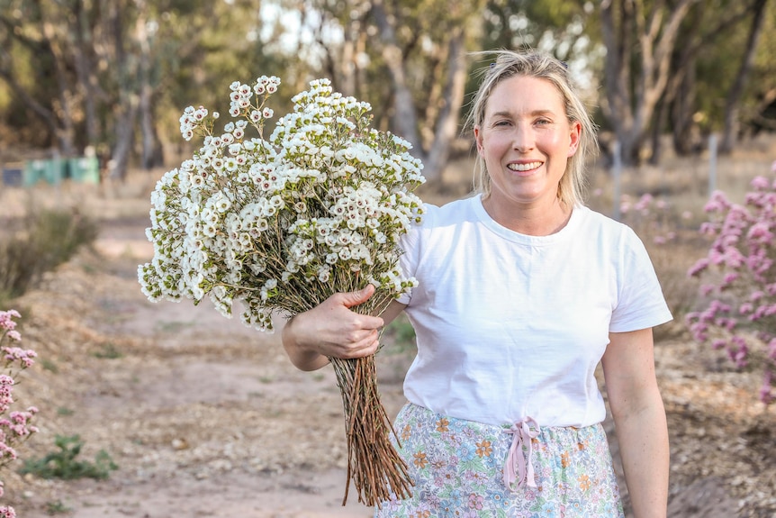 A woman holding a large bunch of white geraldton wax flowers outside with some pink flowers in the background. 