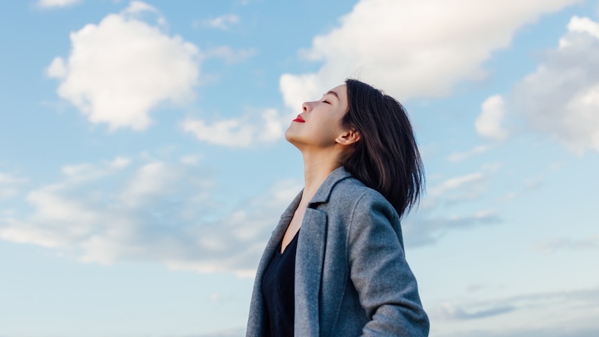 An Asian woman with red lipstick and a grey coat takes a deep breath in through her nose.