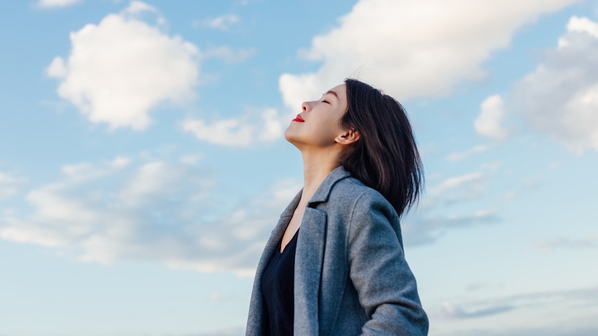 An Asian woman in red lipstick and a gray coat takes a deep breath through her nose.