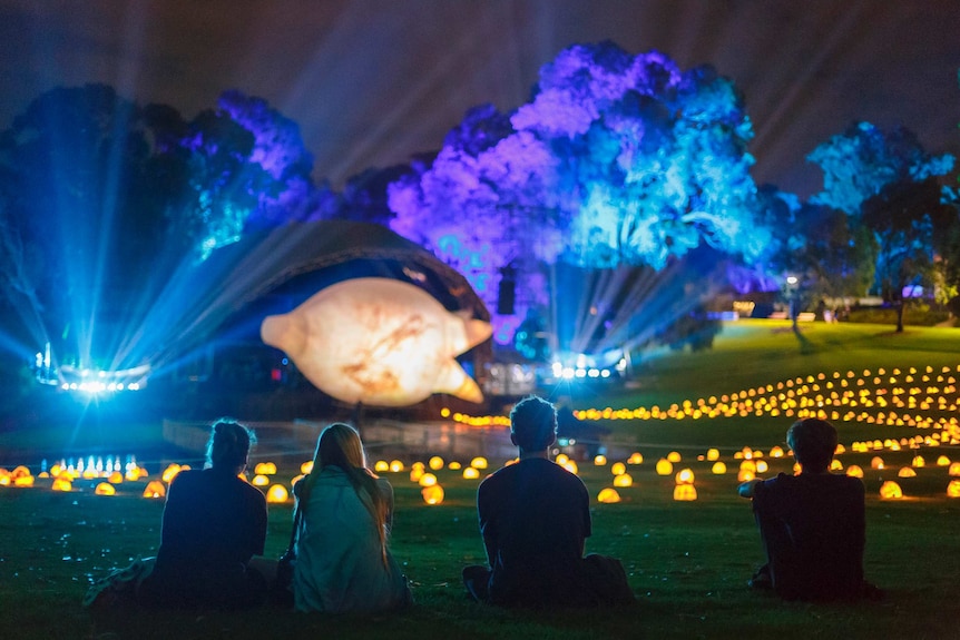 Four people sitting on grass at the Kings Park amphitheatre looking at a colourful light installation.