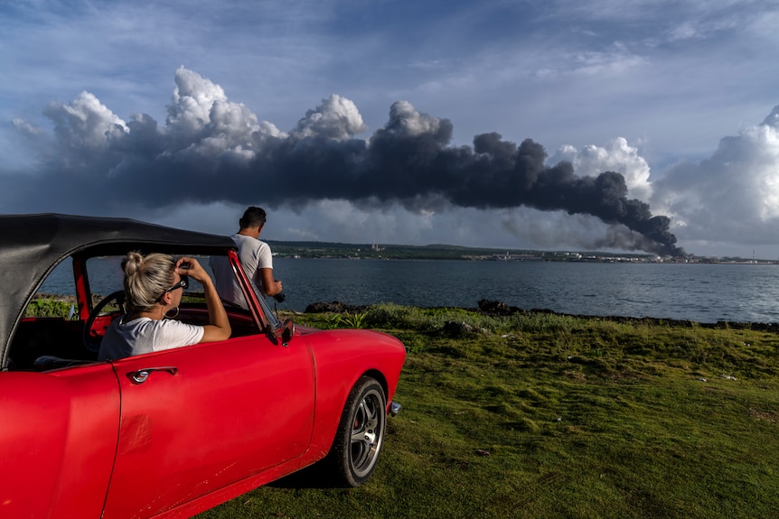 People in a red classic convertible watch smoke rise from across a bay. 