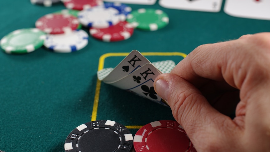 Close-up of a hand lifting two cards with coloured poker chips behind it, on a green felt surface.