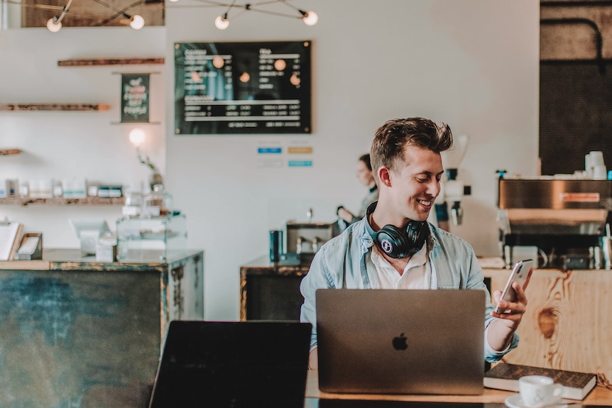 Young man working on laptop in a cafe smiles at his phone 