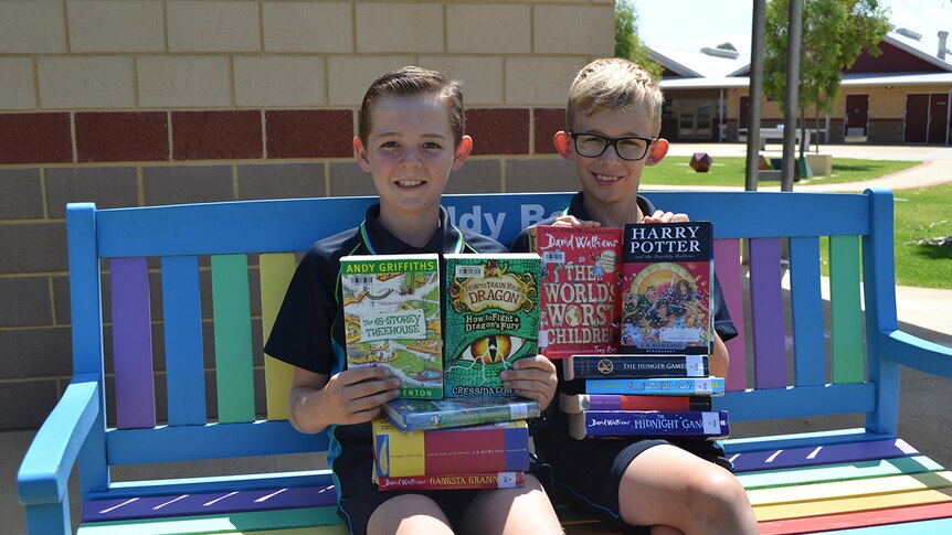 Colour photo of primary school students Matthew and Harper sitting on colourful outdoor bench with stack of books.