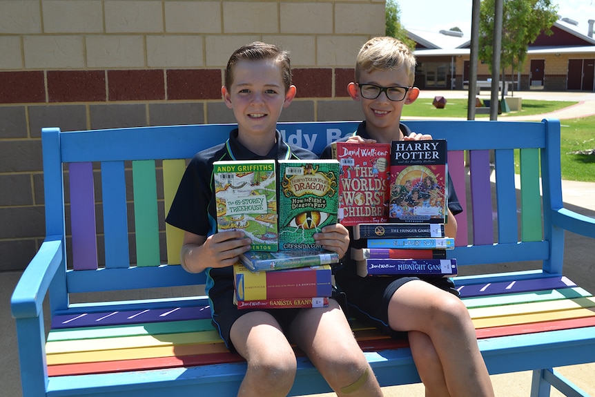 Colour photo of primary school students Matthew and Harper sitting on colourful outdoor bench with stack of books.
