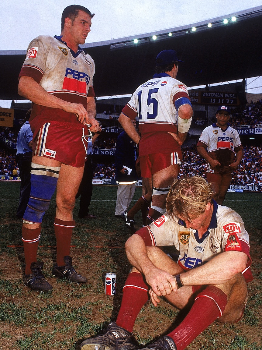 A tall Manly player stands and looks down the field as a teammate sits on the ground with head down after a loss.
