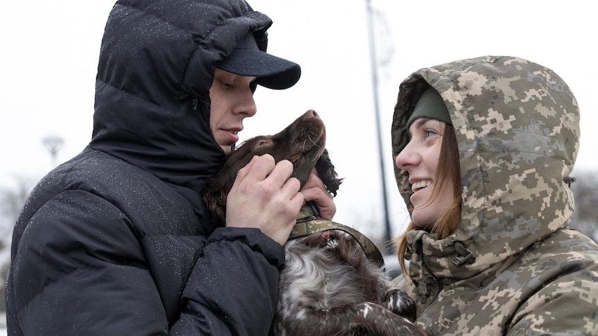 A man in a black hood and a woman in a camo print hood holding a dog. 