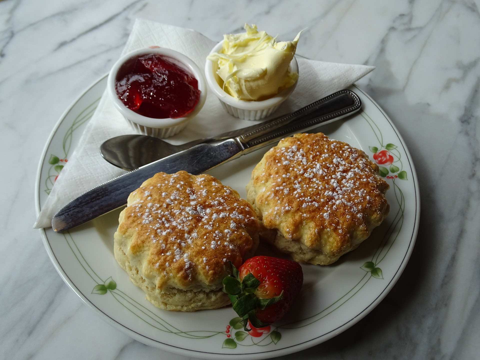Two scones on a plate, with pots of jam and cream, and a fresh strawberry, beside them.
